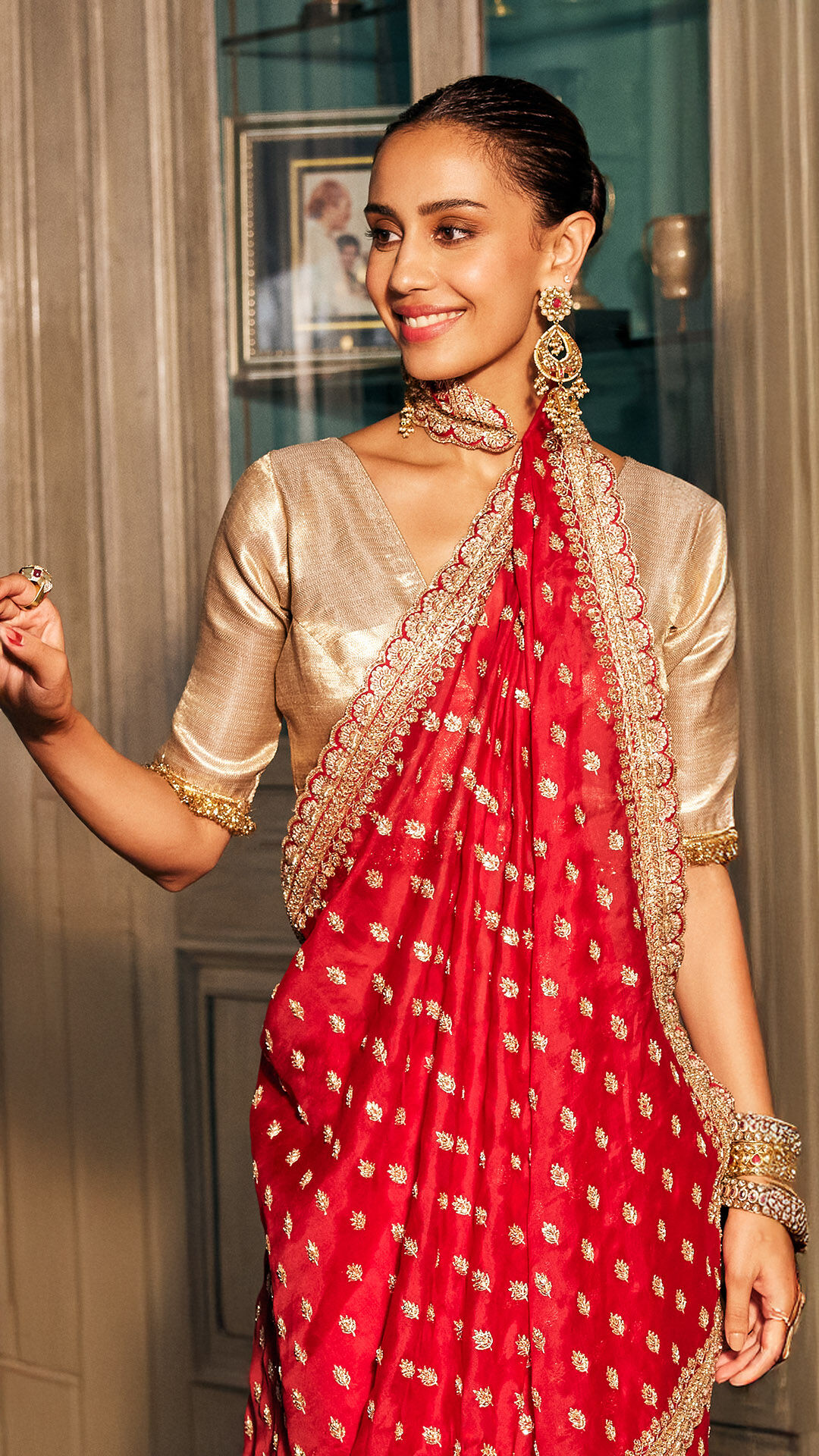 Woman in a traditional red and gold saree with heavy jewelry, smiling gracefully indoors against a classic wooden and glass cabinet backdrop.